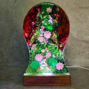 May include: A decorative lamp featuring a miniature koi pond scene encased in clear resin. The lamp showcases koi fish, pink lotus flowers, and green lily pads set against a backdrop of red and green hues. It sits on a wooden base.