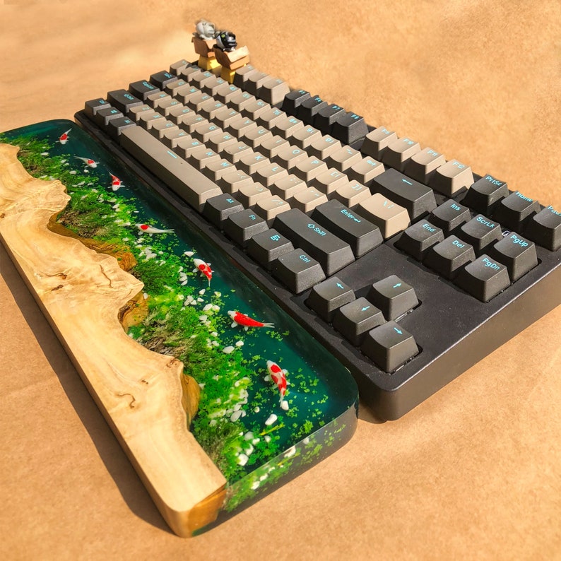 May include: A wooden keyboard wrist rest with a clear epoxy resin top featuring a green and blue landscape with white and red fish. The wrist rest is paired with a black mechanical keyboard with beige keycaps.