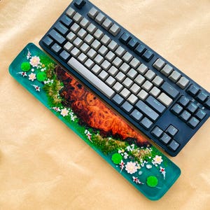 May include: A black mechanical keyboard with gray keycaps rests next to a custom resin wrist rest. The wrist rest features a vibrant design with a wood grain center, surrounded by a teal-colored resin with koi fish, lily pads, and flowers.