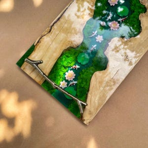 May include: A rectangular wooden serving tray with a river scene. The river is made of blue resin with white and red koi fish, lily pads, and pink and white flowers. The tray has a brass branch handle.