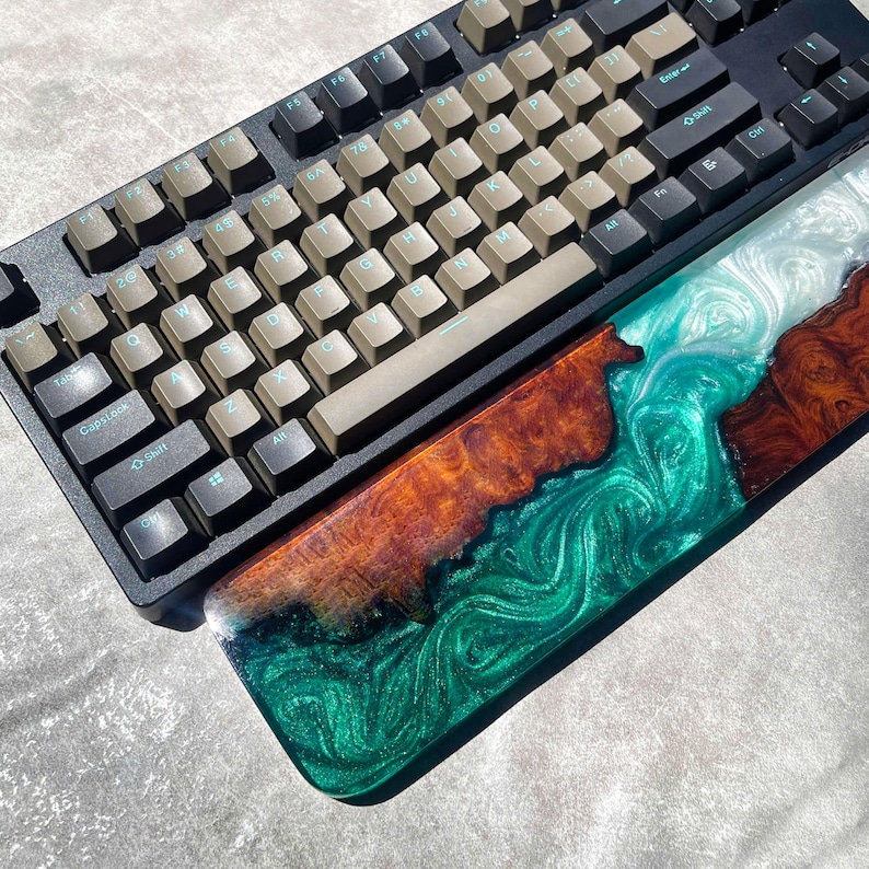 May include: A black mechanical keyboard with tan keycaps and a custom resin wrist rest. The wrist rest features a swirling design with shades of green, white, and brown. The keyboard has a standard layout.