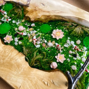Op de afbeelding: Een houten serveerblad met een miniatuur rivierlandschap. De rivier is gevuld met groen mos, kleine stenen en miniatuur koikarpers. Roze lotusbloemen en waterlelies zijn verspreid. De lade heeft een natuurlijke houtnerf afwerking.