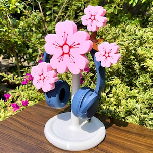 Op de afbeelding: Een witte hoofdtelefoonstandaard met een centrale paal en een ronde voet, bekroond met drie roze kersenbloesemdecoraties. Een donkerblauwe over-ear hoofdtelefoon rust op de standaard, geplaatst onder de roze bloemen.