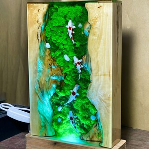 May include: A rectangular wooden lamp with a resin interior featuring a green, moss-like design and white koi fish with red markings. The lamp sits on a wooden base and has a light cord.