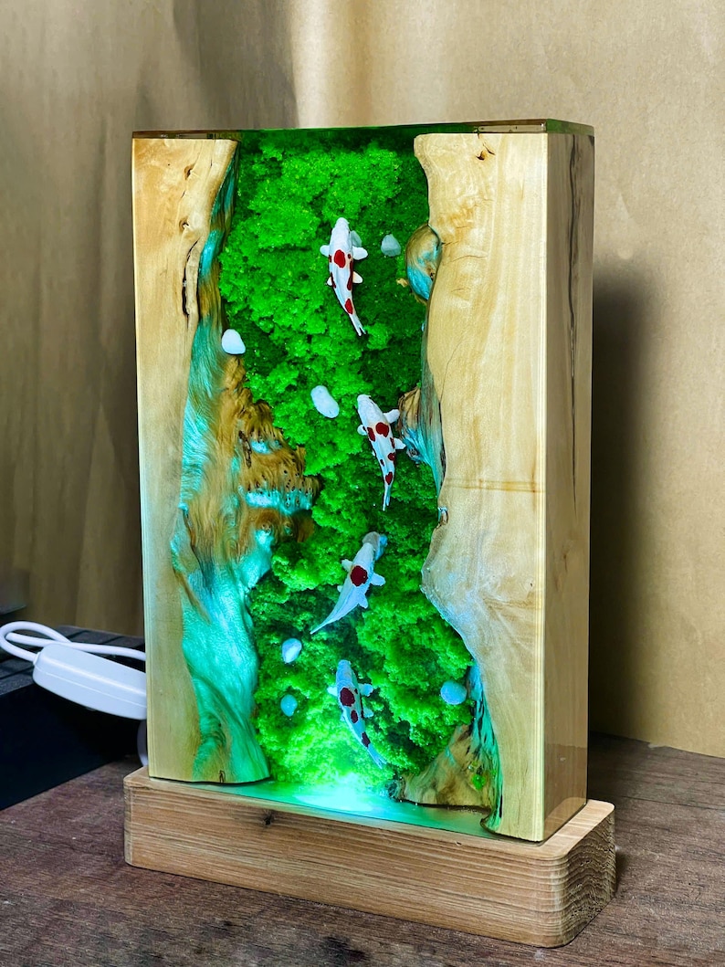 May include: A rectangular wooden lamp with a resin interior featuring a green, moss-like design and white koi fish with red markings. The lamp sits on a wooden base and has a light cord.