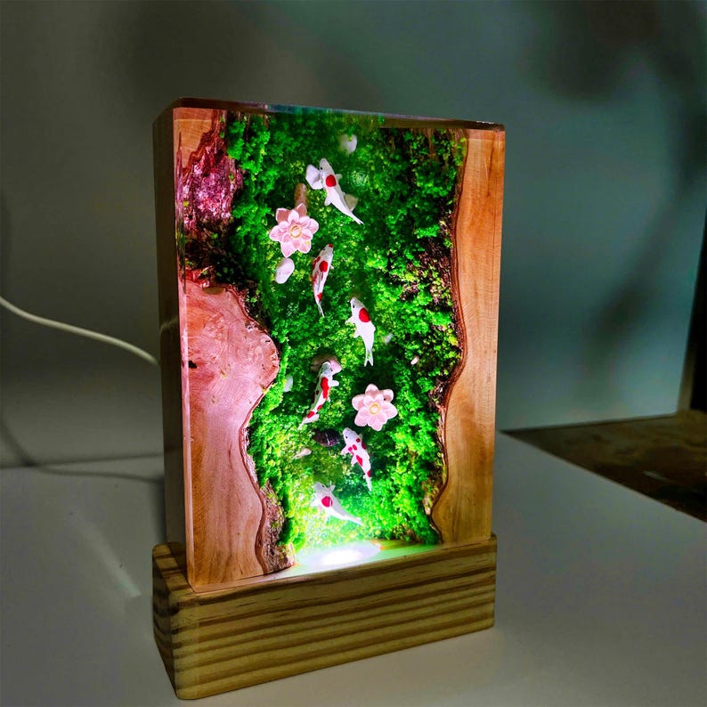 Puede incluir: L&aacute;mpara rectangular de madera con un dise&ntilde;o de resina que presenta peces koi nadando en un paisaje de musgo verde con flores rosas. La l&aacute;mpara se apoya sobre una base de madera y emite un brillo suave.