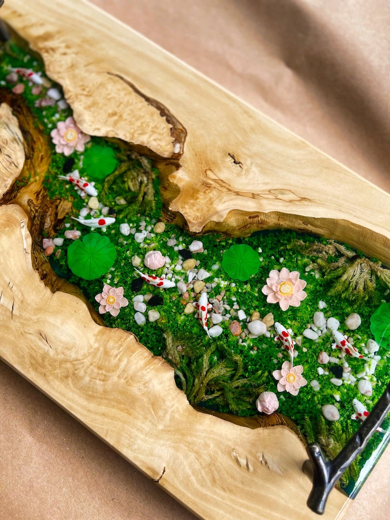 Op de afbeelding: Een rechthoekig houten dienblad met een riviersc&egrave;ne. De rivier is gevuld met groen mos, kleine rotsen en miniatuur koi-vissen. Roze bloemen en waterlelies zijn ook aanwezig. De randen van de lade zijn van natuurlijk hout.