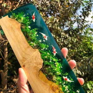 May include: A wooden plaque with a resin inlay depicting a miniature pond scene. The pond is filled with green water and white and red koi fish. The pond is surrounded by green moss and white pebbles.