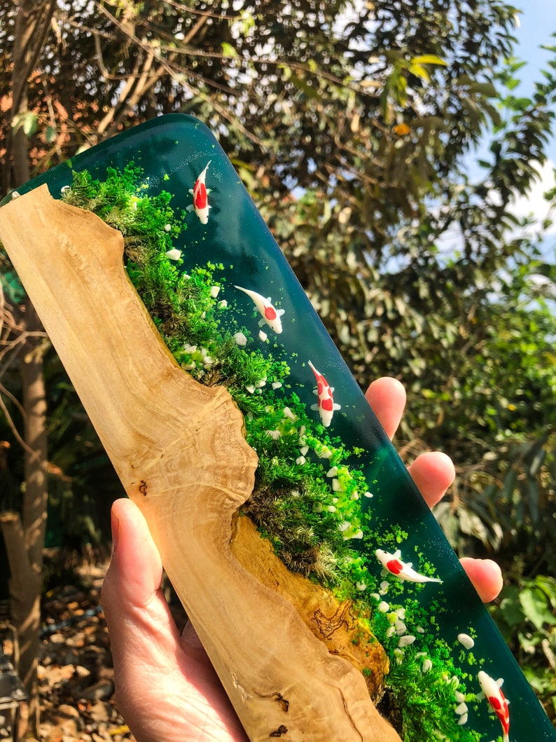 May include: A wooden plaque with a resin inlay depicting a miniature pond scene. The pond is filled with green water and white and red koi fish. The pond is surrounded by green moss and white pebbles.