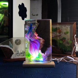 May include: A rectangular resin lamp featuring a vibrant betta fish design with blue and purple hues. The lamp includes a wooden base and is illuminated with colorful LED lights, creating a decorative and artistic lighting piece.