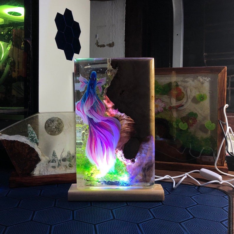 May include: A rectangular resin lamp featuring a vibrant betta fish design with blue and purple hues. The lamp includes a wooden base and is illuminated with colorful LED lights, creating a decorative and artistic lighting piece.