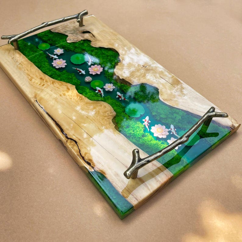 May include: A rectangular wooden serving tray with a river scene. The tray features a green and blue epoxy river with koi fish, lily pads, and flowers. The tray has natural wood edges and bronze branch handles.