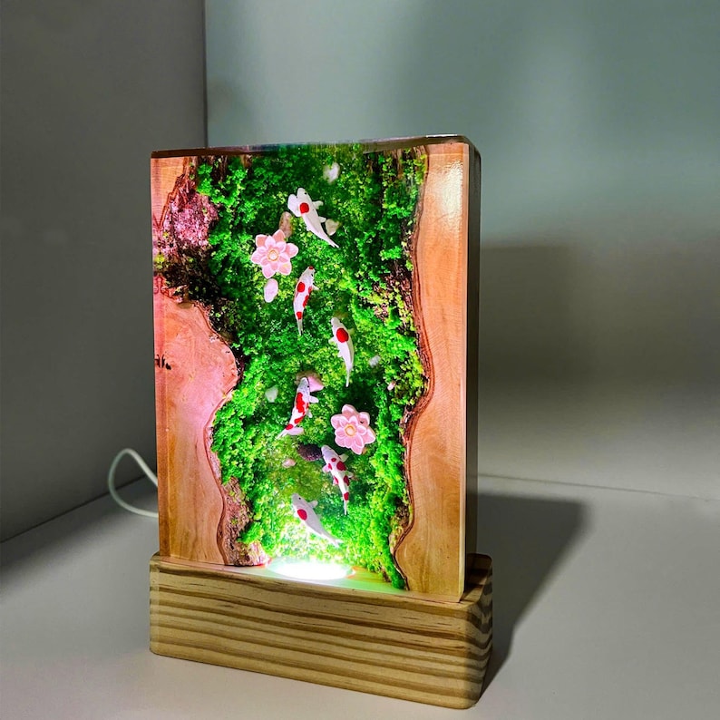 Puede incluir: L&aacute;mpara rectangular de madera con una escena de jard&iacute;n en miniatura con peces koi y flores rosas. La l&aacute;mpara tiene una base de madera y una luz que ilumina la escena desde abajo, creando un brillo c&aacute;lido. Los peces koi son blancos con marcas rojas.