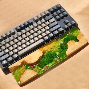 Puede incluir: Un teclado mec&aacute;nico negro con un reposamu&ntilde;ecas de madera que presenta un dise&ntilde;o de resina de musgo verde y peces blancos.