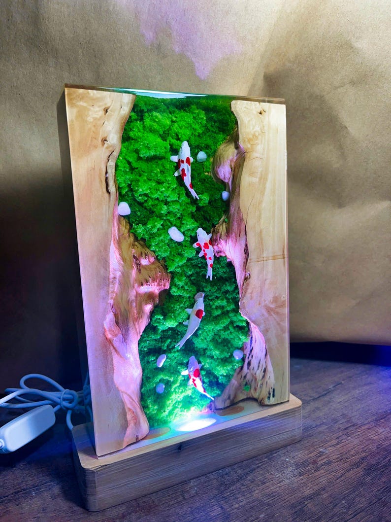 May include: A rectangular wooden lamp with a resin interior featuring a green moss landscape and koi fish. The lamp has a light base and a power cord. The koi fish are white with red markings.