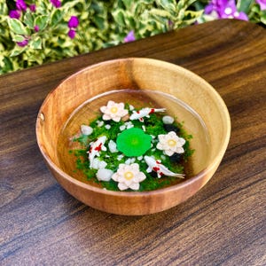 May include: A round wooden bowl filled with clear resin, featuring three miniature white and red koi fish, three pale pink lotus flowers, one bright green lily pad, and small white pebbles embedded in a layer of green moss-like material.