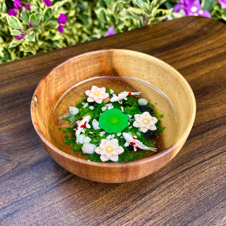 May include: A round wooden bowl filled with clear resin, featuring three miniature white and red koi fish, three pale pink lotus flowers, one bright green lily pad, and small white pebbles embedded in a layer of green moss-like material.
