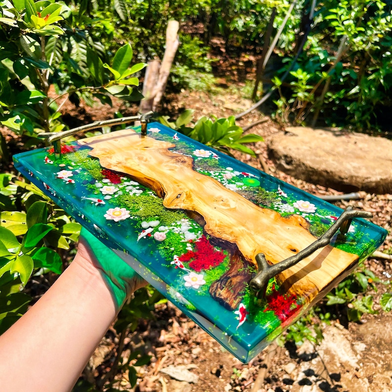 May include: A rectangular serving tray with a clear, turquoise resin base. The tray features a natural wood centerpiece, miniature koi fish, and floral accents. The tray has bronze-colored handles.