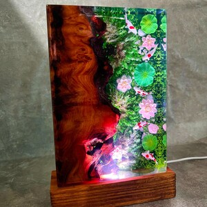 May include: A rectangular wooden lamp with a unique design. The lamp features a wooden base and a clear resin panel. The resin panel showcases a vibrant scene with koi fish, pink flowers, and green lily pads. The wood has a rich, reddish-brown color.