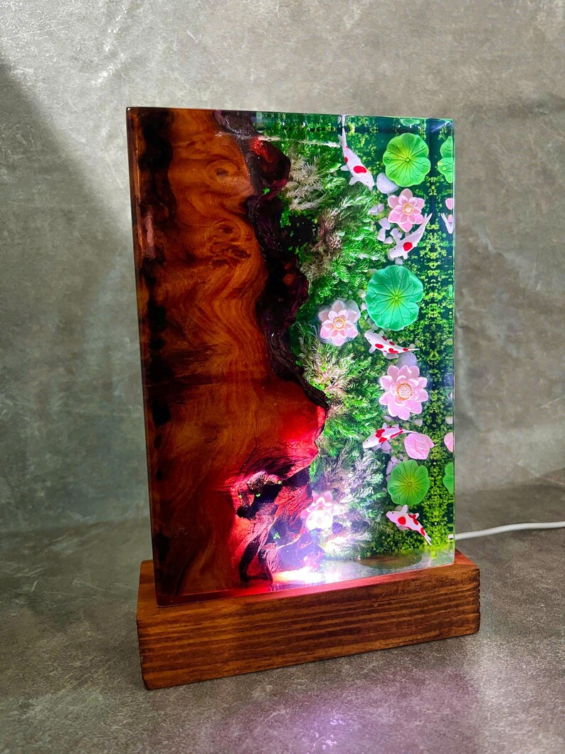 May include: A rectangular wooden lamp with a unique design. The lamp features a wooden base and a clear resin panel. The resin panel showcases a vibrant scene with koi fish, pink flowers, and green lily pads. The wood has a rich, reddish-brown color.