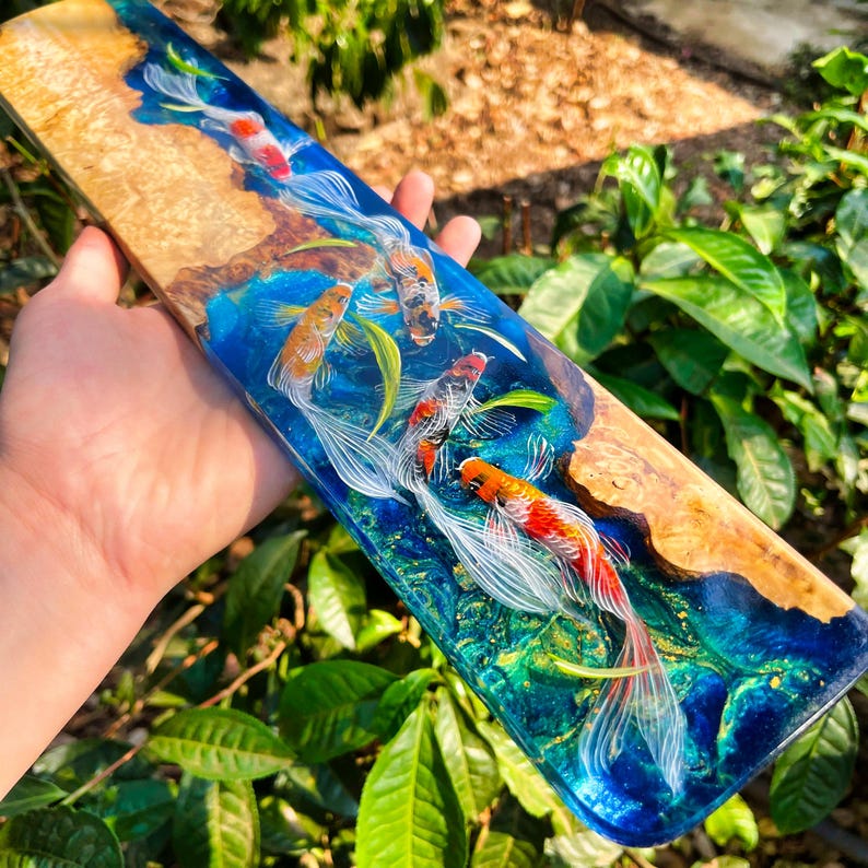 May include: A rectangular wooden serving board with a vibrant epoxy resin design. The board features a natural wood grain on one side and a blue and green resin river with painted koi fish. The koi are orange, red, and white.