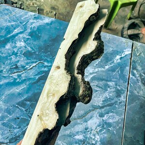 Puede incluir: Una tabla de madera clara y sin tratar con un canal profundo, org&aacute;nico y marr&oacute;n oscuro tallado en su centro. La veta y la textura naturales de la madera son visibles, mostrando su car&aacute;cter &uacute;nico. La tabla est&aacute; inclinada contra una superficie azul similar al m&aacute;rmol.