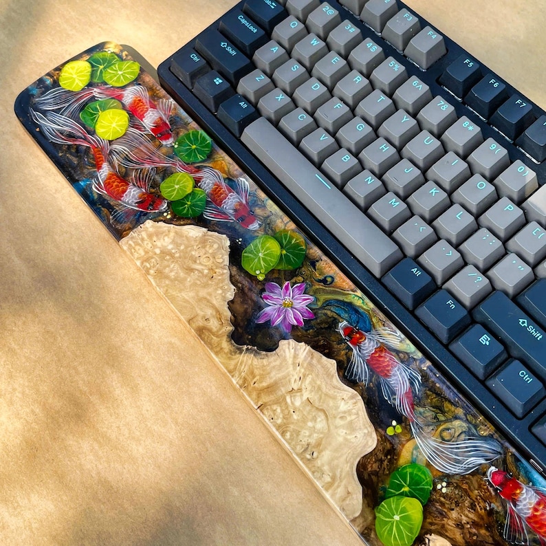 May include: A custom keyboard with a wrist rest featuring a koi pond design. The wrist rest is made of wood and resin, with a detailed painting of koi fish, lily pads, and a lotus flower. The keyboard has gray and black keycaps.
