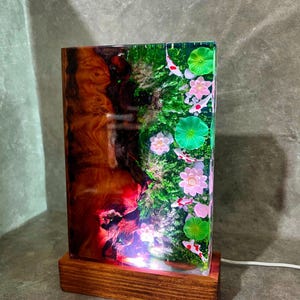 May include: A rectangular resin lamp with a wooden base. The lamp features a koi pond scene with pink flowers, green lily pads, and koi fish. The other side has a wood grain design with red and brown hues.