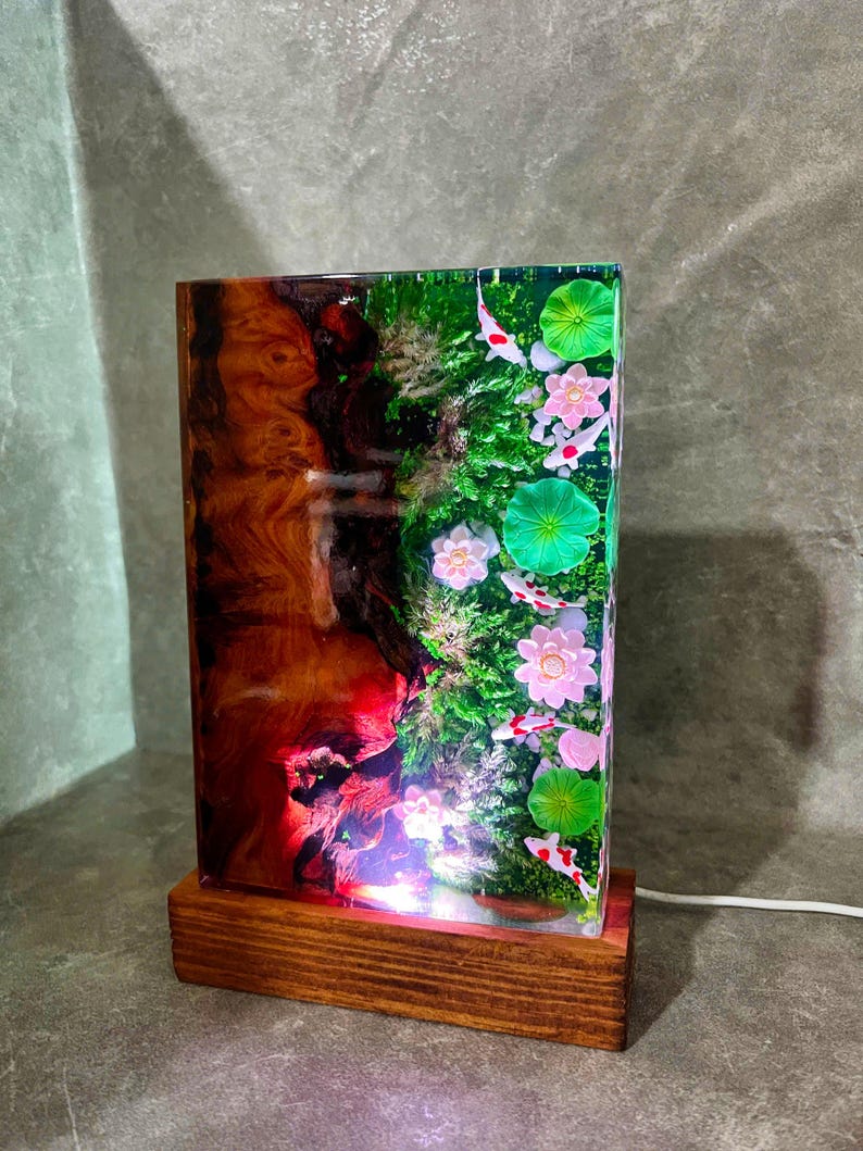 May include: A rectangular resin lamp with a wooden base. The lamp features a koi pond scene with pink flowers, green lily pads, and koi fish. The other side has a wood grain design with red and brown hues.