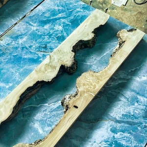 Puede incluir: Dos tablas de madera con bordes naturales, con tonos marr&oacute;n claro y beige con acentos de corteza marr&oacute;n oscuro. Las tablas est&aacute;n colocadas sobre una superficie azul con aspecto de m&aacute;rmol, mostrando la veta y textura &uacute;nicas de la madera.