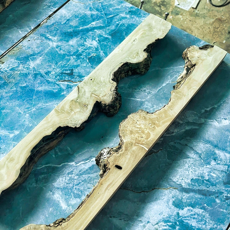 Puede incluir: Dos tablas de madera con bordes naturales, con tonos marr&oacute;n claro y beige con acentos de corteza marr&oacute;n oscuro. Las tablas est&aacute;n colocadas sobre una superficie azul con aspecto de m&aacute;rmol, mostrando la veta y textura &uacute;nicas de la madera.