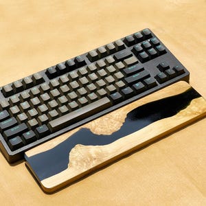 K&ouml;nnte beinhalten: Eine schwarze mechanische Tastatur mit hellgr&uuml;ner Beschriftung liegt &uuml;ber einer h&ouml;lzernen Handgelenkauflage. Die Handgelenkauflage zeigt einen Fluss aus schwarzem Harz, der durch helles Holz flie&szlig;t und ein einzigartiges Design schafft.