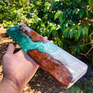 May include: A rectangular wooden and resin art piece. The piece features a combination of brown wood, swirling white, and teal resin. The wood has a natural grain pattern. The resin has a glossy finish. The piece is held in a hand.