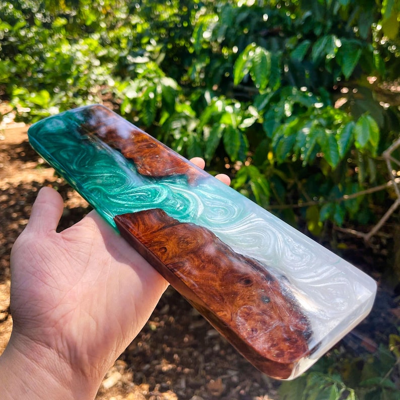 May include: A rectangular wooden and resin art piece. The piece features a combination of brown wood, swirling white, and teal resin. The wood has a natural grain pattern. The resin has a glossy finish. The piece is held in a hand.