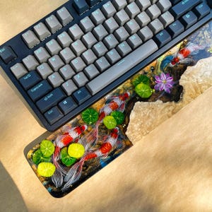 May include: A black mechanical keyboard with gray keycaps rests above a custom wrist rest. The wrist rest features a detailed resin design of koi fish, lily pads, and a purple flower, set against a wood grain background.