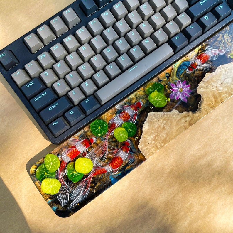May include: A black mechanical keyboard with gray keycaps rests above a custom wrist rest. The wrist rest features a detailed resin design of koi fish, lily pads, and a purple flower, set against a wood grain background.