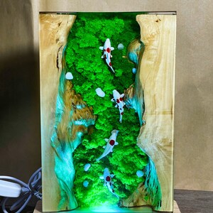May include: A rectangular wooden lamp with a clear resin center featuring a green moss landscape and several koi fish with red and black markings. The lamp sits on a wooden base and has a power cord.