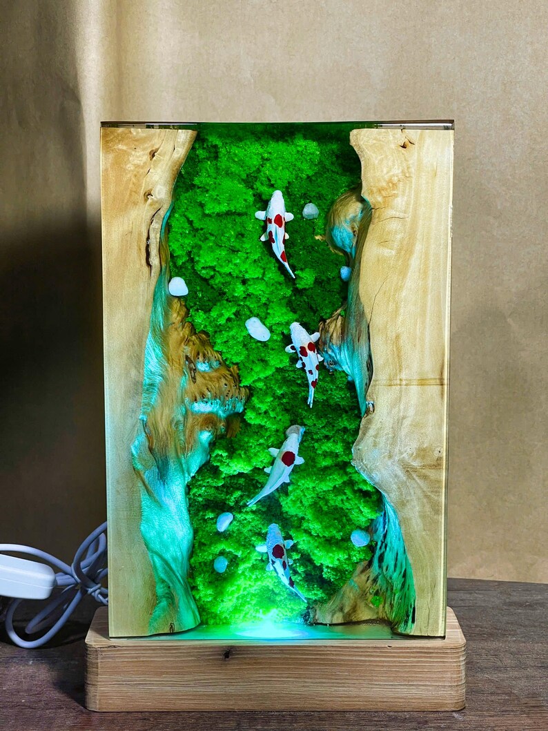 May include: A rectangular wooden lamp with a clear resin center featuring a green moss landscape and several koi fish with red and black markings. The lamp sits on a wooden base and has a power cord.