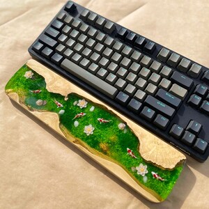 Puede incluir: Un teclado mec&aacute;nico negro con teclas grises descansa sobre un reposamu&ntilde;ecas de madera. El reposamu&ntilde;ecas presenta un dise&ntilde;o de r&iacute;o de resina epoxi verde con peces koi, flores blancas y peque&ntilde;as rocas.