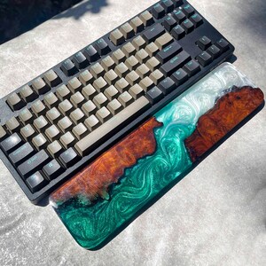 May include: A black mechanical keyboard with tan keycaps sits above a custom wrist rest. The wrist rest features a wood grain design with green and white epoxy resin. The keyboard and wrist rest are on a gray surface.