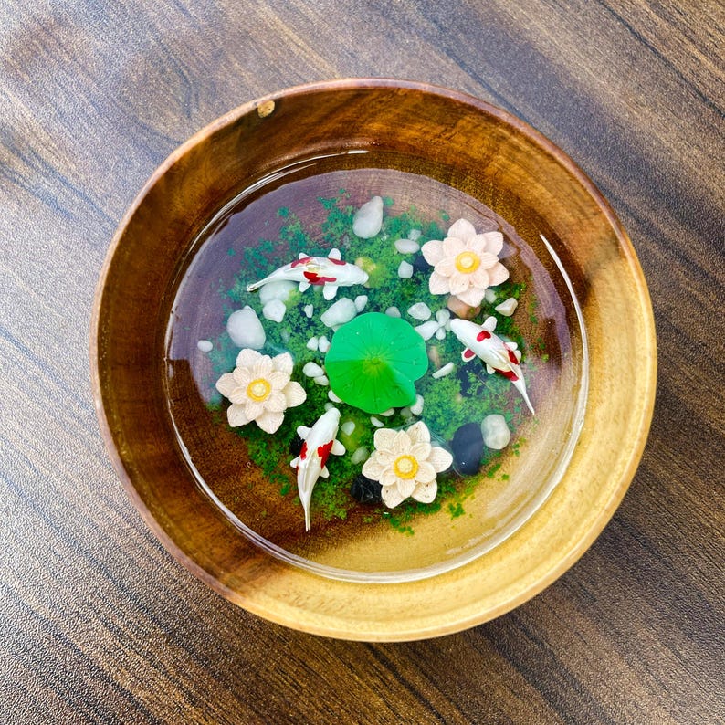 May include: A round wooden bowl filled with clear resin containing three miniature white and red koi fish, three pale pink lotus flowers, a single bright green lily pad, and small white and black pebbles scattered over a green mossy base.