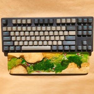Puede incluir: Un teclado mec&aacute;nico negro con un juego de teclas de color gris claro y un reposamu&ntilde;ecas de madera con un dise&ntilde;o de musgo verde y peces.