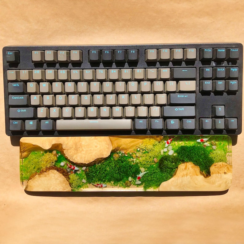 Puede incluir: Un teclado mec&aacute;nico negro con un juego de teclas de color gris claro y un reposamu&ntilde;ecas de madera con un dise&ntilde;o de musgo verde y peces.