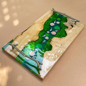 May include: A rectangular wooden serving tray with a river design. The river is filled with green moss, lily pads, and koi fish. Pink and white flowers are scattered throughout the river. The tray has metal branch handles.