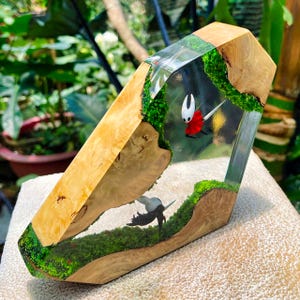 May include: A wooden art piece with a clear resin center, featuring a miniature scene with moss and cartoon characters. The wood has a light brown color with darker patterns. The resin section shows a red and white character.