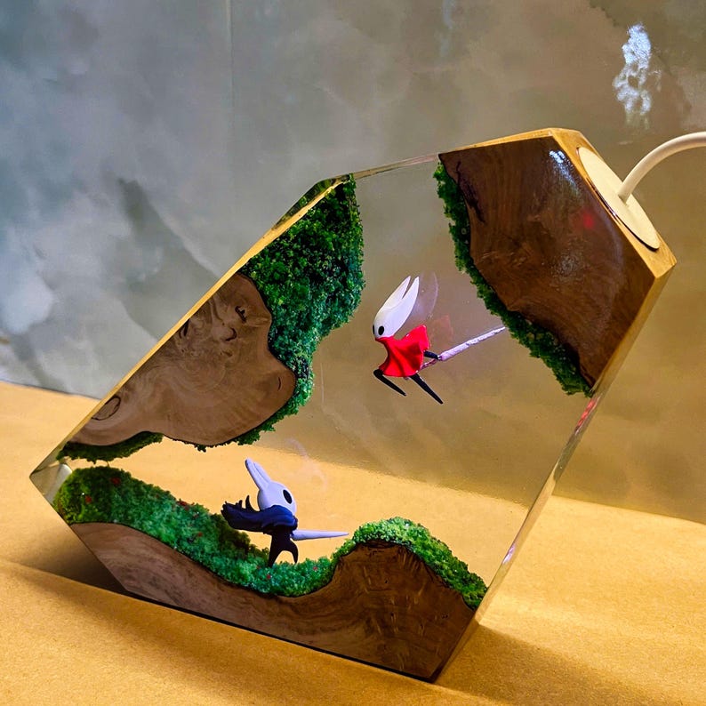 May include: A unique resin art piece featuring two stylized figures in a dynamic pose, set within a landscape of green moss and wooden elements. The figures are in red, white, and black. The piece has a wooden frame and a white cord.