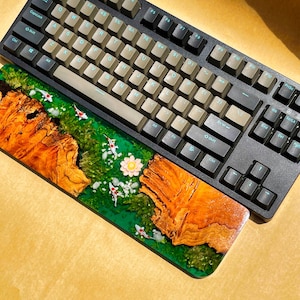 May include: A black mechanical keyboard with tan keycaps rests above a custom resin wrist rest. The wrist rest features a vibrant design with koi fish, flowers, and a wood grain pattern set in green resin. The keyboard has a standard layout.