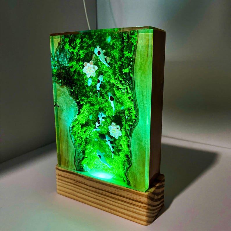 Puede incluir: L&aacute;mpara rectangular de madera con interior verde brillante, con dise&ntilde;o de peces koi y flores blancas. La l&aacute;mpara se apoya en una base de madera y emite una suave luz verde.