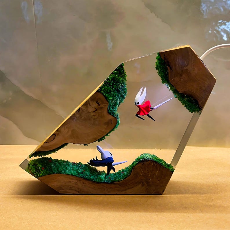 May include: A unique resin art piece featuring two stylized figures in a dynamic pose within a clear, geometric form. The figures are set against a backdrop of green moss and wooden elements, creating a miniature landscape.