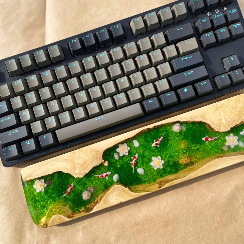 Puede incluir: Un teclado mec&aacute;nico con teclas grises y un reposamu&ntilde;ecas de madera personalizado. El reposamu&ntilde;ecas presenta un dise&ntilde;o de r&iacute;o de resina epoxi verde con peces koi, flores blancas y peque&ntilde;as piedras.